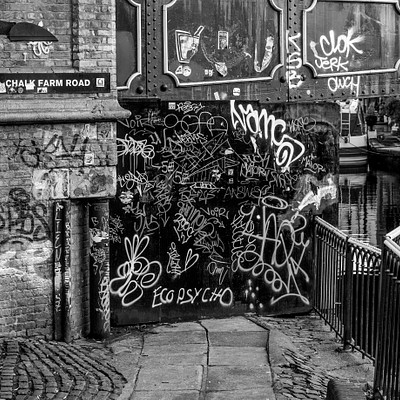 The image is a black and white photo of an alleyway with graffiti on the walls. There are several signs posted along the alley, including one that reads "Schwinn Road." A bridge can be seen in the background, adding to the urban atmosphere of the scene. In addition to the graffiti, there is a boat visible near the top right corner of the image.