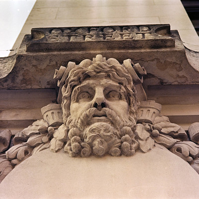 The image features a large stone building with an ornate design on the front. A prominent gargoyle statue is prominently displayed, adding to the architectural beauty of the structure. The gargoyle has a beard and appears to be looking downward, giving it a menacing appearance.