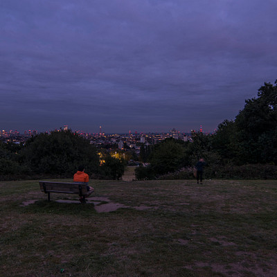 The image features a park with a bench situated in the middle of it. A person is sitting on the bench, enjoying the view of the city skyline in the distance. There are several other people scattered throughout the park, some closer to the bench and others further away.