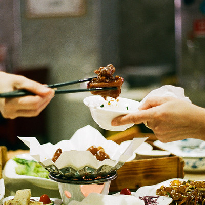 The image depicts a group of people gathered around a dining table, enjoying a meal together. There are several bowls filled with food placed on the table, and some individuals are using chopsticks to eat their meals. A person is holding up a plate of food for another person to take a bite from it.