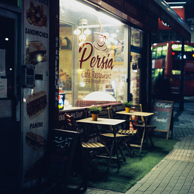 The image features a restaurant with a Persia theme, located on the side of a street. There are several tables and chairs arranged outside the establishment, creating an inviting atmosphere for customers to enjoy their meals. A bus is parked nearby, adding to the urban setting.