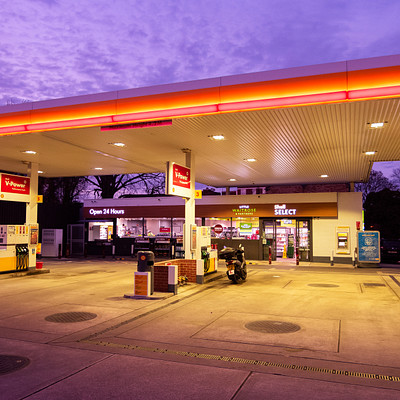 The image features a gas station with a large Shell sign on top. There are several cars parked in the lot, including one near the center of the scene and another towards the right side. A motorcycle is also visible, parked closer to the left side of the lot.