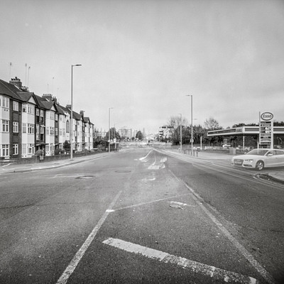 The image is a black and white photo of an empty street in the middle of town. There are several cars parked along the sides of the road, with one car on each side of the street. A few people can be seen walking or standing near the parked cars.