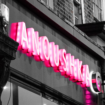 The image features a large pink neon sign on the side of a building, which reads "Anoushka." The sign is illuminated and stands out against the dark background. There are also two smaller signs visible in the scene, one located near the top left corner and another towards the bottom right corner.