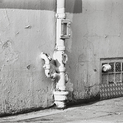 The image is a black and white photo of an old fire hydrant on the sidewalk. It appears to be rusted, indicating its age. The fire hydrant is located next to a building with a pipe sticking out from it.