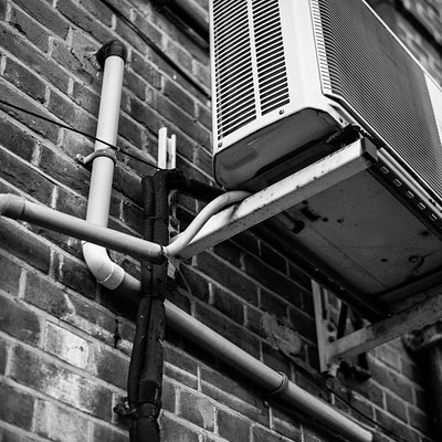 The image features a black and white photo of an air conditioning unit mounted on the side of a brick wall. The air conditioner is positioned above a pipe, which runs along the wall. There are also two pipes visible in the scene, one located near the top left corner and another at the bottom right corner.