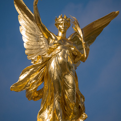 The image features a large, golden statue of an angel or goddess. The statue is positioned on top of a pedestal and appears to be made of gold or brass. It has wings spread out, giving it the appearance of flying through the air.