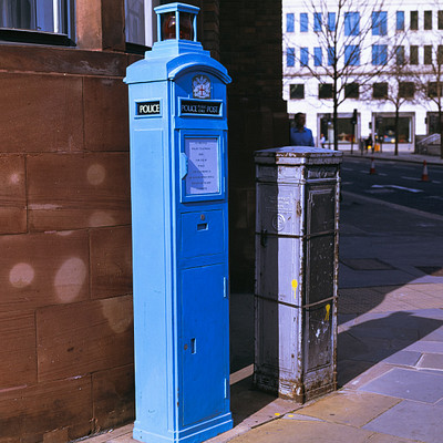 The image features a blue mailbox sitting on the sidewalk next to a building. It is positioned in front of a brick wall, and there are two other mailboxes nearby. One of these mailboxes is located closer to the left edge of the scene, while the other one is situated further back on the right side.