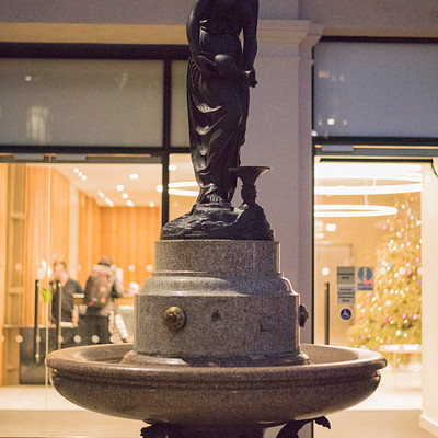 The image features a statue of a woman holding a child, standing on top of a fountain. The statue is positioned in front of a building with a window nearby. There are several people around the area, some closer to the statue and others further away.