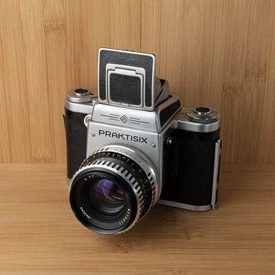 The image features an old-fashioned camera sitting on a wooden table. The camera is silver and black in color, with its lens facing the viewer. It appears to be an antique model, possibly a Praktisix, which adds a sense of nostalgia to the scene.