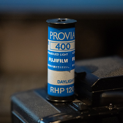 The image features a blue film canister with the word "Provia" written on it. It is placed in front of a computer monitor, which occupies most of the background. The film canister appears to be Fuji Film branded and has a 400 speed.