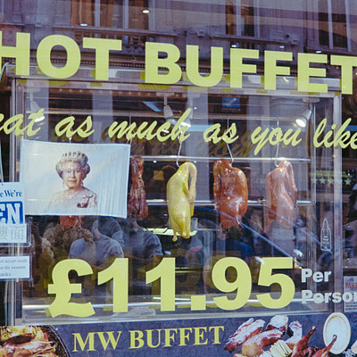 The image features a restaurant window with an advertisement for Hot Buffet. The sign is displayed prominently in the window, and it appears to be a place where people can enjoy various foods as much as they like. Inside the restaurant, there are several people present, some of whom seem to be enjoying their meals or conversing with one another.