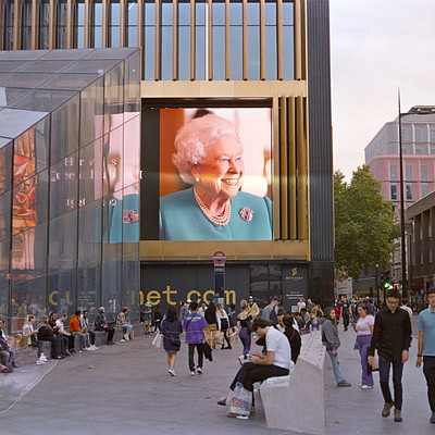 The image depicts a busy city street with people walking around and sitting on benches. There are several individuals scattered throughout the scene, some of them carrying handbags or backpacks. A large billboard featuring Queen Elizabeth is visible in the background, drawing attention from passersby.