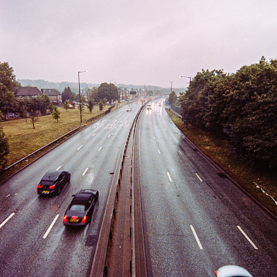 The image is a black and white photo of a highway with several cars driving on it. There are at least five cars visible in the scene, some closer to the foreground while others are further away. A truck can also be seen among the vehicles.