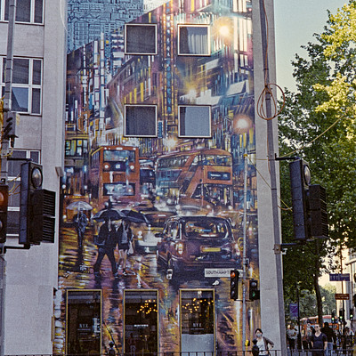 The image features a large building with a mural painted on its side. The mural is of a busy city street, complete with cars and people. There are several traffic lights visible in the scene, including one near the top left corner, another at the bottom right corner, and two more towards the center-right area.