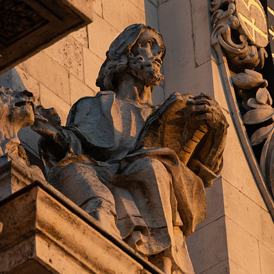 The image features a statue of an old man sitting on a bench, holding a book in his hands. He appears to be reading the book while seated. The statue is located near a building with a clock mounted on its side.