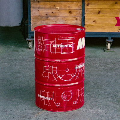 The image features a red metal barrel with white lettering on it, sitting in the middle of a parking lot. The barrel is positioned near a wooden building and appears to be an industrial-sized container. There are also two trucks visible in the background, one located towards the right side of the image and another further back on the left side.