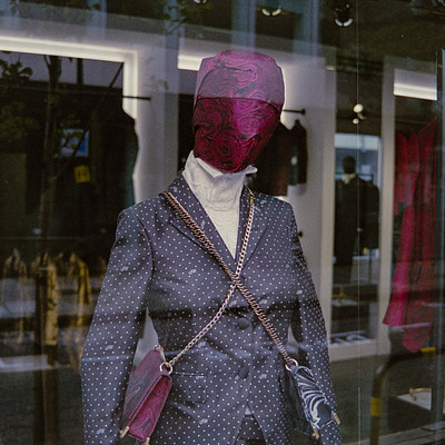 The image features a mannequin dressed in a suit and tie, wearing a red mask. The mannequin is standing in front of a window display with clothes hanging around it. There are several other people visible in the background, possibly browsing or shopping for clothing items.