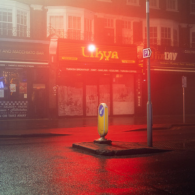 The image depicts a rainy night in an urban setting, with a red traffic light illuminating the street. A yellow parking meter is situated on the sidewalk near the corner of the street. There are several people walking around, some closer to the camera and others further away.