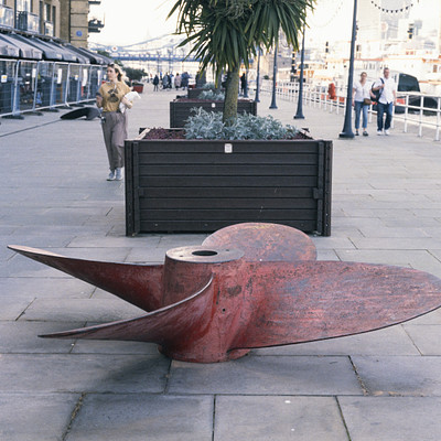 The image features a large red sculpture of an airplane wing, placed on the sidewalk. It is situated in front of a building and near some benches. There are several people walking around the area, with one person standing close to the sculpture and others scattered throughout the scene.