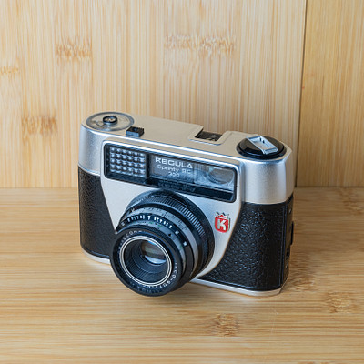 The image features a vintage camera sitting on top of a wooden table. The camera is an old-fashioned model, possibly a Kodak, and it has a leather case around it. The camera appears to be well-maintained and ready for use.
