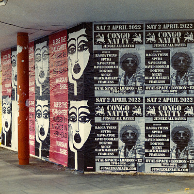 The image is a black and white photo of a wall with various posters on it. There are several posters featuring faces, including one large face that takes up a significant portion of the wall. Other smaller faces can be seen in different areas of the wall as well.