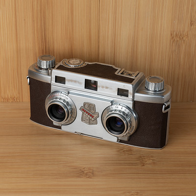 The image features an old-fashioned camera sitting on a wooden table. The camera is silver and brown in color, with two lenses visible on the front. It appears to be an antique or vintage model, possibly from the 1950s era.