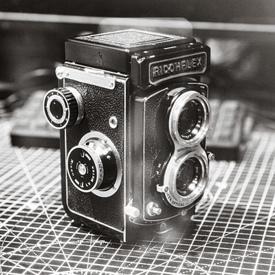 The image is a black and white photograph of an old-fashioned camera. It appears to be a vintage model, possibly from the 1950s or earlier. The camera has a leather case around it, adding to its antique charm.