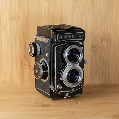 The image features a vintage black and white camera sitting on top of a wooden table. The camera is an old-fashioned model, possibly a Ricohflex, with two lenses visible in the front. It appears to be a classic piece of photography equipment that has been preserved or displayed for its historical value.