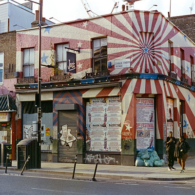 The image features a colorful building with a large mural on its side, painted in red and blue. There are several people walking around the area outside of the building, including two women who appear to be crossing the street. In addition to the pedestrians, there is a car parked nearby, as well as a couple of trucks visible in the scene.