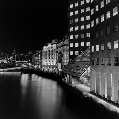 The image is a black and white photo of a city at night, featuring a large building with many windows. A boat can be seen on the water in front of the building, adding to the urban atmosphere. There are also several cars parked around the area, further emphasizing the bustling city environment.