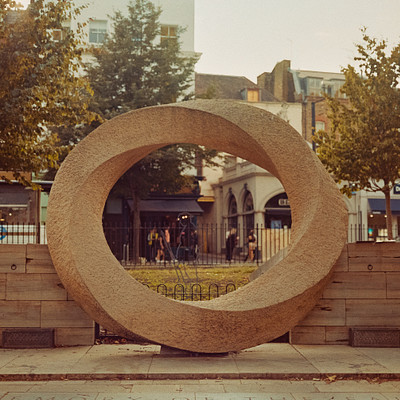 The image features a large stone circle, or cement ring, located in the middle of a city street. This unique structure is surrounded by trees and benches, providing a peaceful spot for people to sit and relax. Several individuals can be seen walking around the area, enjoying the atmosphere created by this artistic installation.