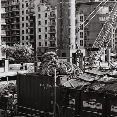 The image is a black and white photo of a large crane in the middle of a city. The crane is surrounded by several buildings, including some tall ones that are visible in the background. There are also two trucks parked nearby, one on the left side and another further back to the right.