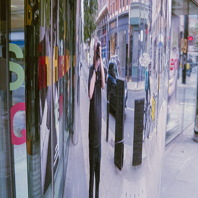 The image is a reflection of a city street scene, captured through the glass window of a building. In the reflection, there are several people walking on the sidewalk and cars parked along the street. A person can be seen taking a picture of themselves in the mirror, capturing their reflection as well.