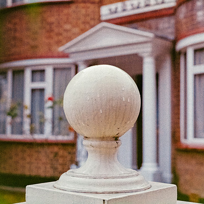 The image features a white ball sitting on top of a white pedestal, which is located in front of a house. The house has a red brick exterior and appears to be an old-style building with columns. There are several windows visible throughout the scene, some of which have curtains drawn over them.