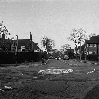 The image is a black and white photo of an intersection in a residential area. There are several cars parked on the street, with some located near the center of the scene and others closer to the edges. A few people can be seen walking around or standing at various points within the scene.