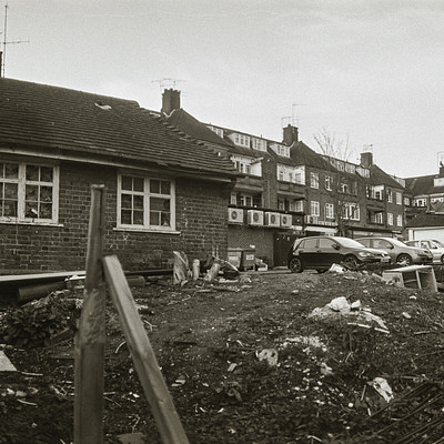 The image is a black and white photo of an old house with a fenced-in yard. There are several cars parked outside the house, including one on the left side, two in the middle, and another on the right side. A truck can also be seen parked near the center of the scene.