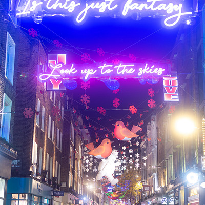 The image depicts a bustling city street at night, illuminated by colorful lights and decorations. There are numerous people walking around the area, with some carrying handbags and backpacks. A large sign is visible above the crowd, adding to the festive atmosphere of the scene.