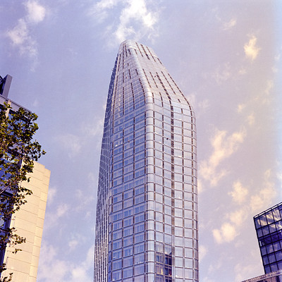 The image features a tall, modern building with many windows. It is situated in front of other buildings and appears to be the focal point of the scene. The building's design resembles an antenna or a large glass spire, giving it a unique appearance.