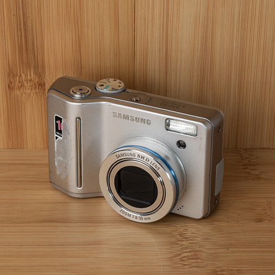 The image features a silver Samsung digital camera sitting on top of a wooden table. The camera is placed in the center of the scene, with its front facing towards the viewer. The wooden surface provides an appealing backdrop for this modern device.