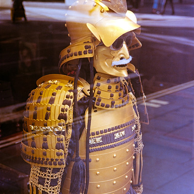 The image features a statue of a samurai warrior, dressed in traditional armor and holding a sword. The statue is positioned on the sidewalk near a street, possibly as an art display or decoration. In addition to the main statue, there are several other people visible in the scene, some walking by and others standing nearby.