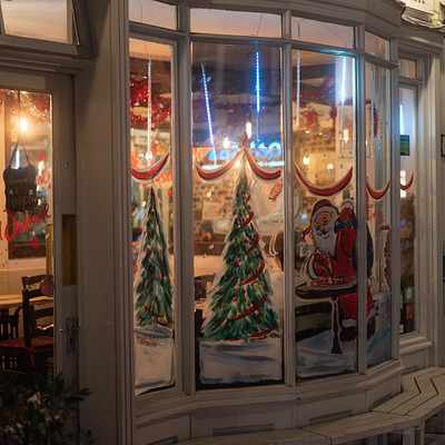 The image features a storefront with a large window display that showcases Christmas trees and Santa Claus. There are several chairs placed around the area, likely for customers to sit while they shop or enjoy their time in the store. A dining table can also be seen within the scene, adding to the cozy atmosphere of the establishment.