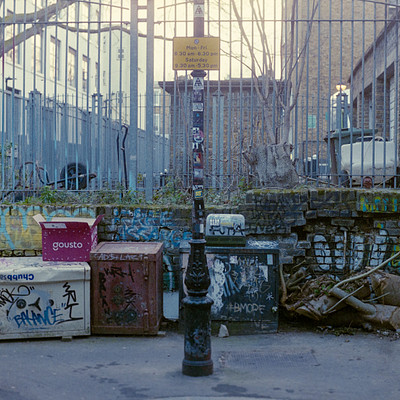 The image depicts a fenced-in area with several boxes and trash cans. There are three large cardboard boxes placed in the middle of the scene, while two smaller trash cans are located closer to the right side of the area. A pole is situated near the center of the scene, and there's also a fire hydrant on the left side.