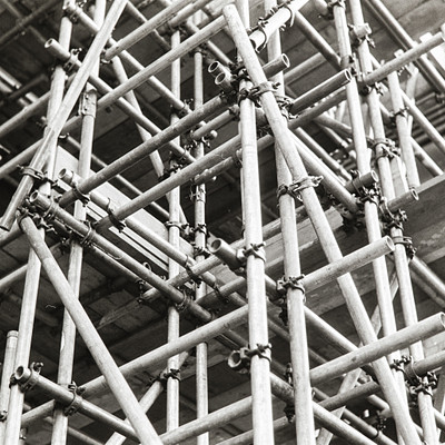 The image is a black and white photo of a large construction site with many wooden scaffolding beams. These beams are arranged in various stacks, creating an impressive structure that dominates the scene. There are numerous metal brackets attached to these wooden beams, providing additional support for the construction project.