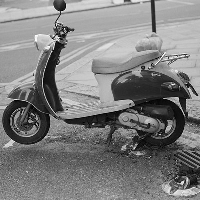 The image is a black and white photo of an old motor scooter parked on the sidewalk. It appears to be broken down, as it has fallen over onto its side. The scooter is located near a curb, with a fire hydrant visible in the background.