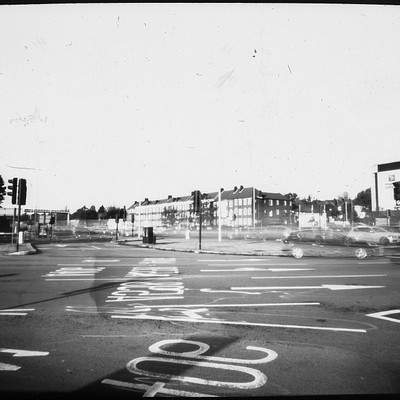 The image is a black and white photo of an empty street intersection with several cars parked on the side. There are multiple traffic lights in the scene, some closer to the foreground while others are further away. A stop sign can also be seen near the left edge of the frame.