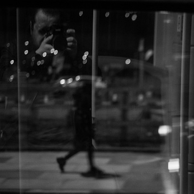 The image is a black and white photo of a man taking a picture of himself in the mirror. He appears to be using a camera, capturing his reflection as he stands on a sidewalk. There are several cars parked nearby, with one car closer to the left edge of the frame and two others further back on the right side.