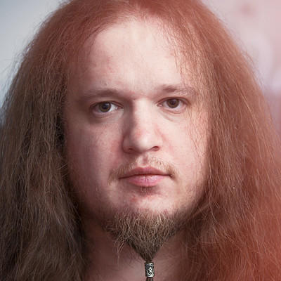 The image features a man with long, red hair and a beard. He has a goatee and is wearing a necklace around his neck. His eyes are open wide as he stares at the camera.