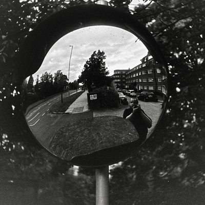 The image is a black and white photo of a street scene, taken through the reflection in a mirror. In the mirror's reflection, there are several cars parked along the side of the road, as well as a few people walking or standing around. A traffic light can be seen on one side of the street, adding to the urban atmosphere.