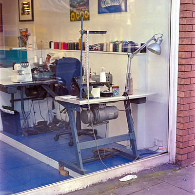 The image is a black and white photo of a cleaning business with a window display. Inside the shop, there are several sewing machines arranged on tables, along with various office supplies such as chairs, desks, and books. A large table occupies most of the space in the room, while other smaller tables can be seen throughout the area.